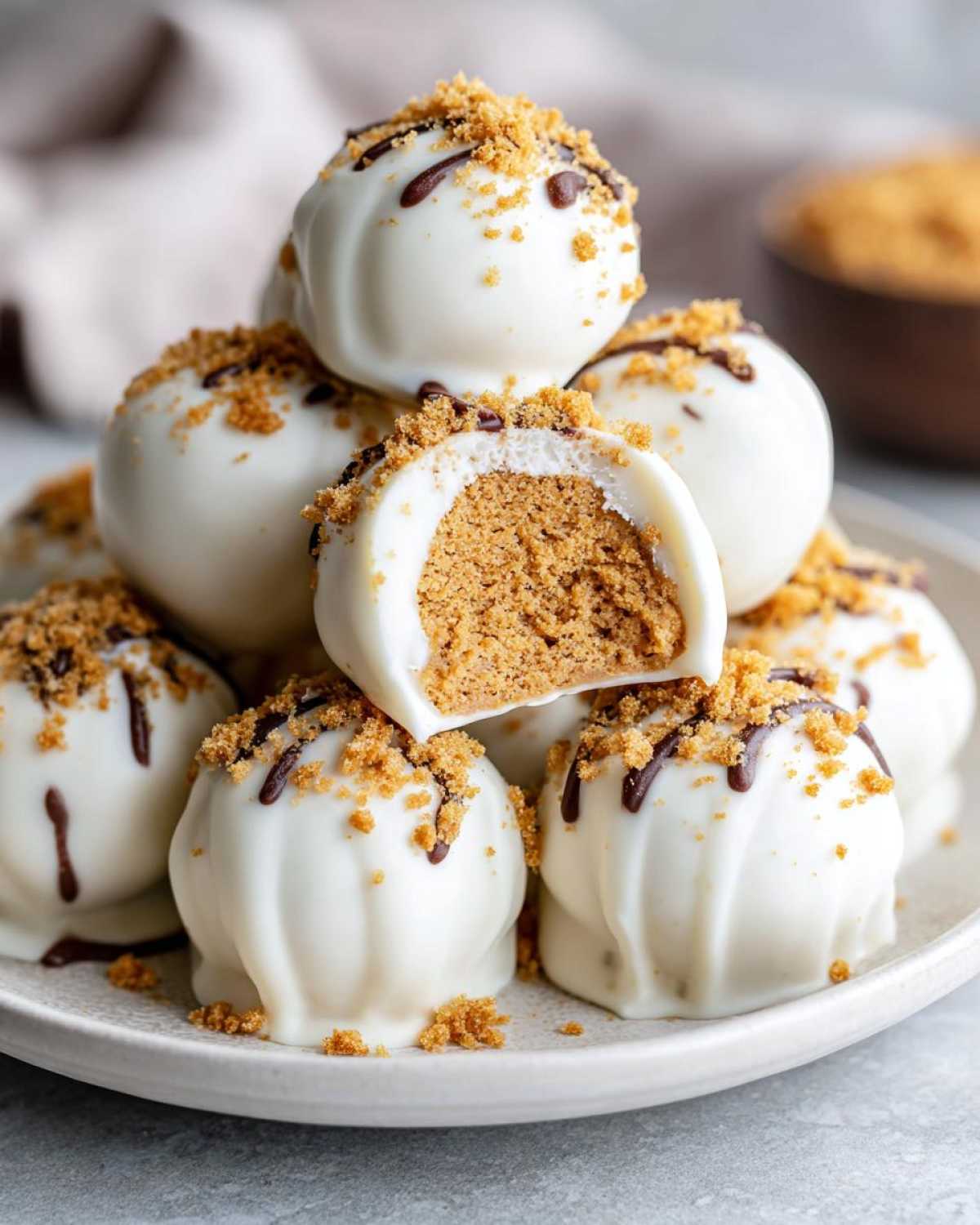 A stack of white chocolate covered pumpkin cheesecake truffles, one cut in half to show the creamy interior, topped with graham cracker crumbs and chocolate drizzle.