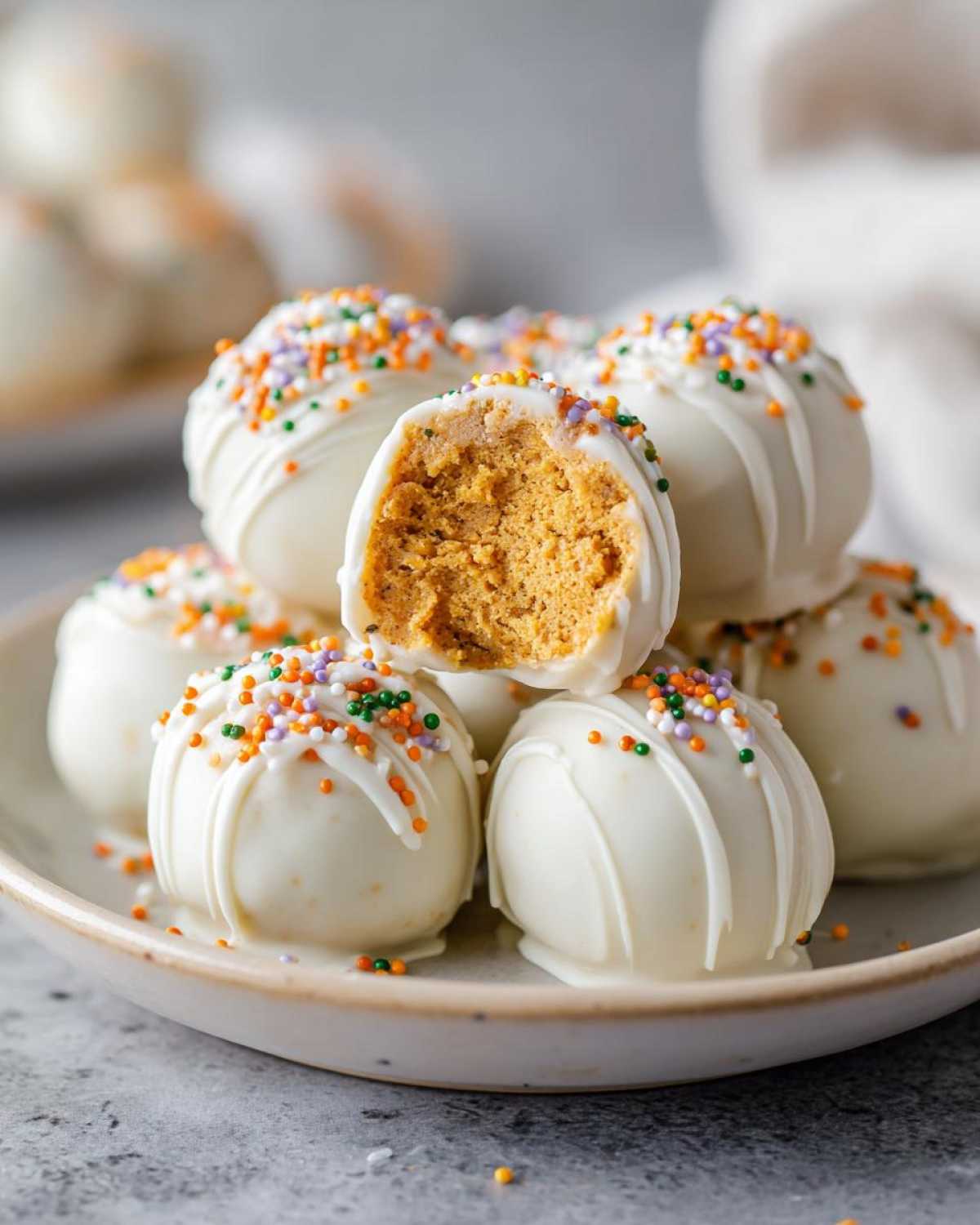A stack of white chocolate-covered Pumpkin Oreo Balls, one bitten into, showing the pumpkin filling and festive sprinkles.