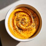 A swirl of vibrant orange Pumpkin Whipped Feta Dip in a white bowl, topped with herbs and spices.