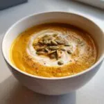 A bowl of creamy slow cooker pumpkin soup topped with a swirl of cream, pumpkin seeds, and spices.