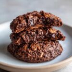 Close-up of a stack of three rich, dark chocolate almond joy cookies with visible coconut flakes and melted chocolate chips.