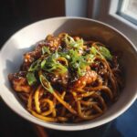 A close-up of a bowl filled with glossy sticky garlic chicken noodles, topped with sesame seeds and chopped green onions.