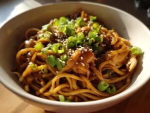 A close-up of a bowl of sticky garlic chicken noodles, garnished with green onions and sesame seeds.