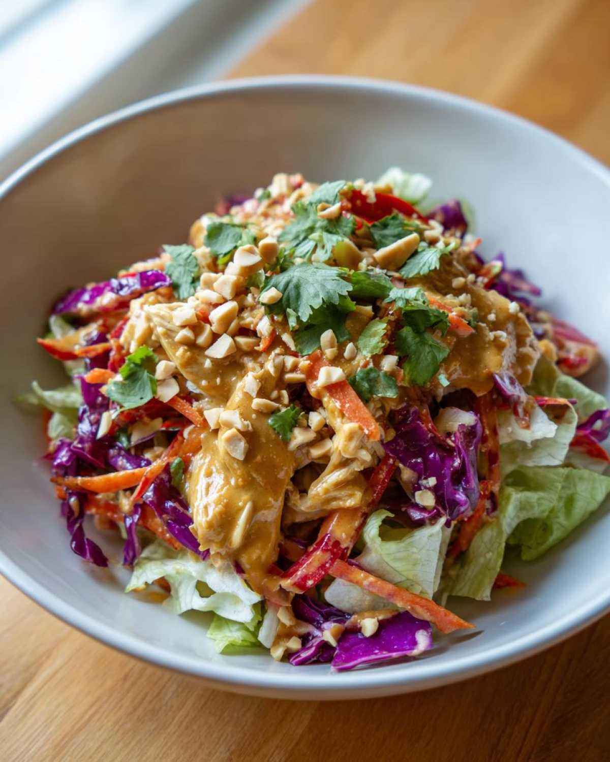Close-up of a vibrant thai chicken salad in a white bowl, topped with peanut sauce, chopped peanuts, and cilantro.
