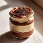 A layered tiramisu chia pudding in a glass jar, topped with cocoa powder.