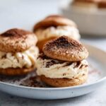Close-up of two tiramisu cookies stacked with a creamy filling and dusted with cocoa powder.