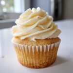 A single, beautifully frosted vanilla cupcake with creamy white buttercream sitting on a white surface.