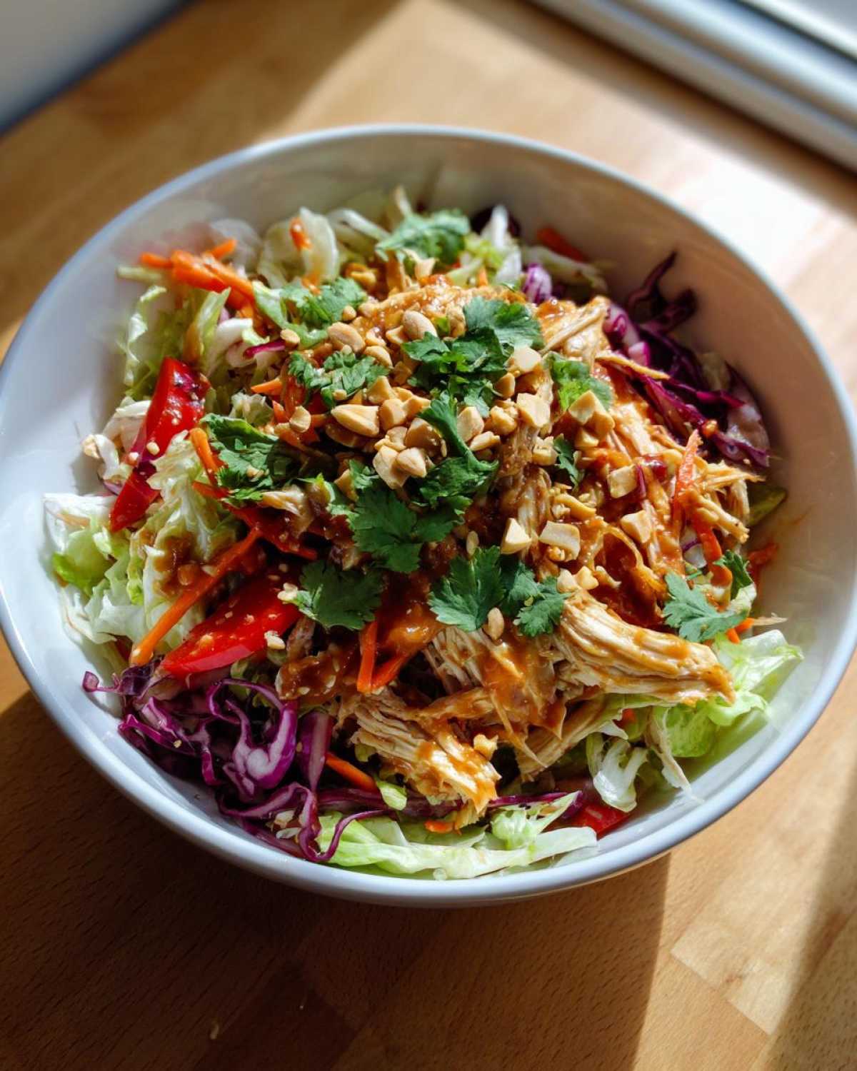 A vibrant bowl of thai chicken salad featuring shredded chicken, mixed greens, red cabbage, carrots, and topped with peanuts and cilantro.
