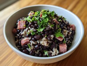 A close-up of a white bowl filled with flavorful black beans and rice mixed with chunks of ham and topped with fresh cilantro.