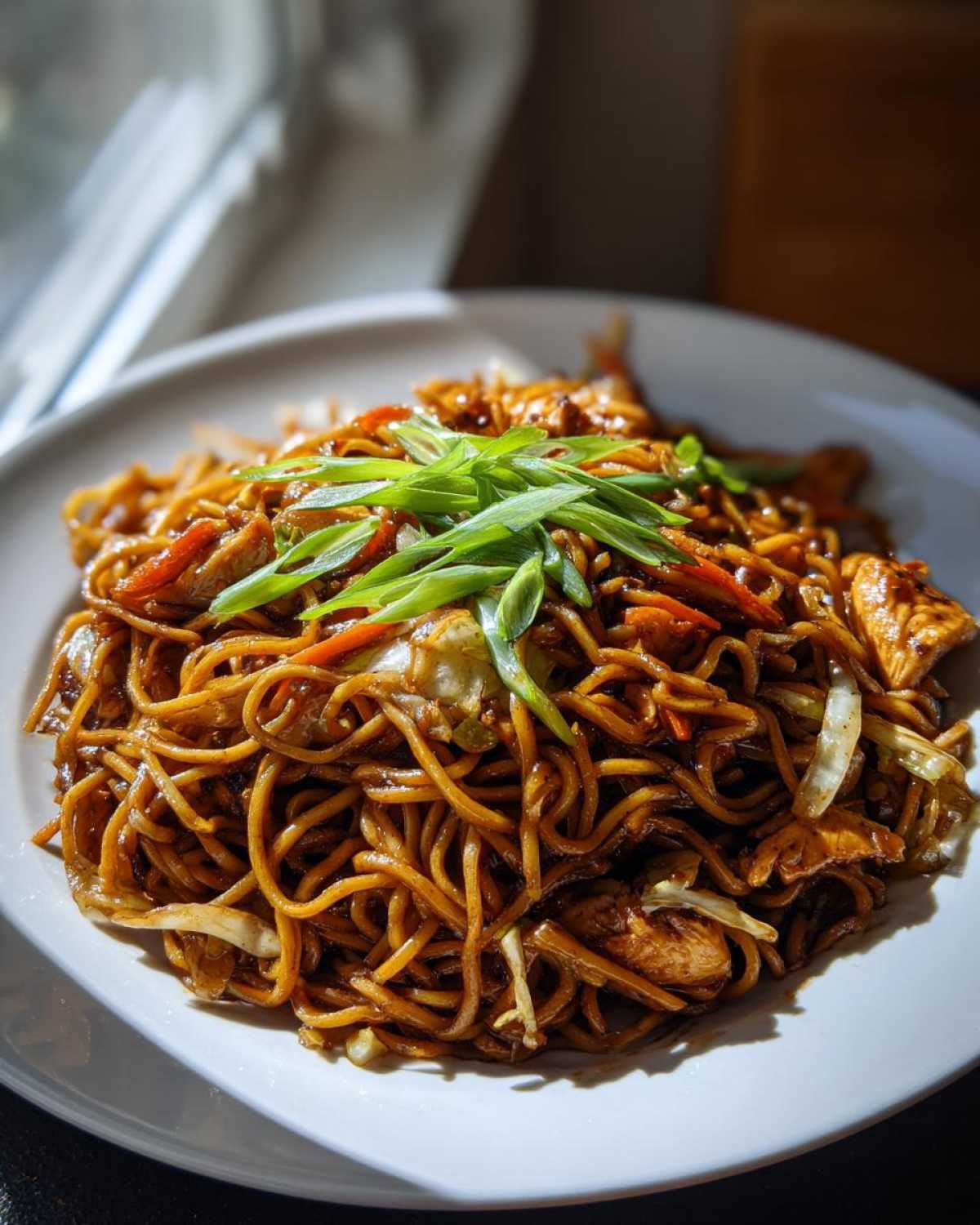 Close-up of a plate piled high with glossy, dark brown chicken chow mein recipe noodles, topped with fresh green onions.