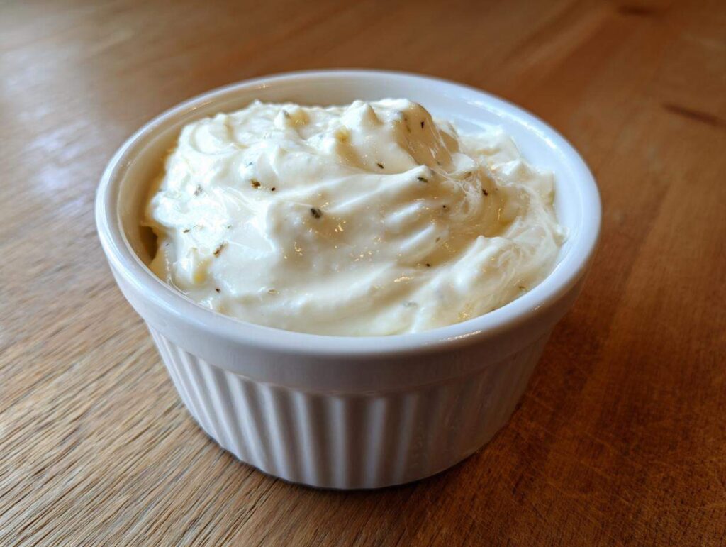 A close-up of thick, creamy garlic sauce speckled with herbs in a small white ramekin on a wooden surface.