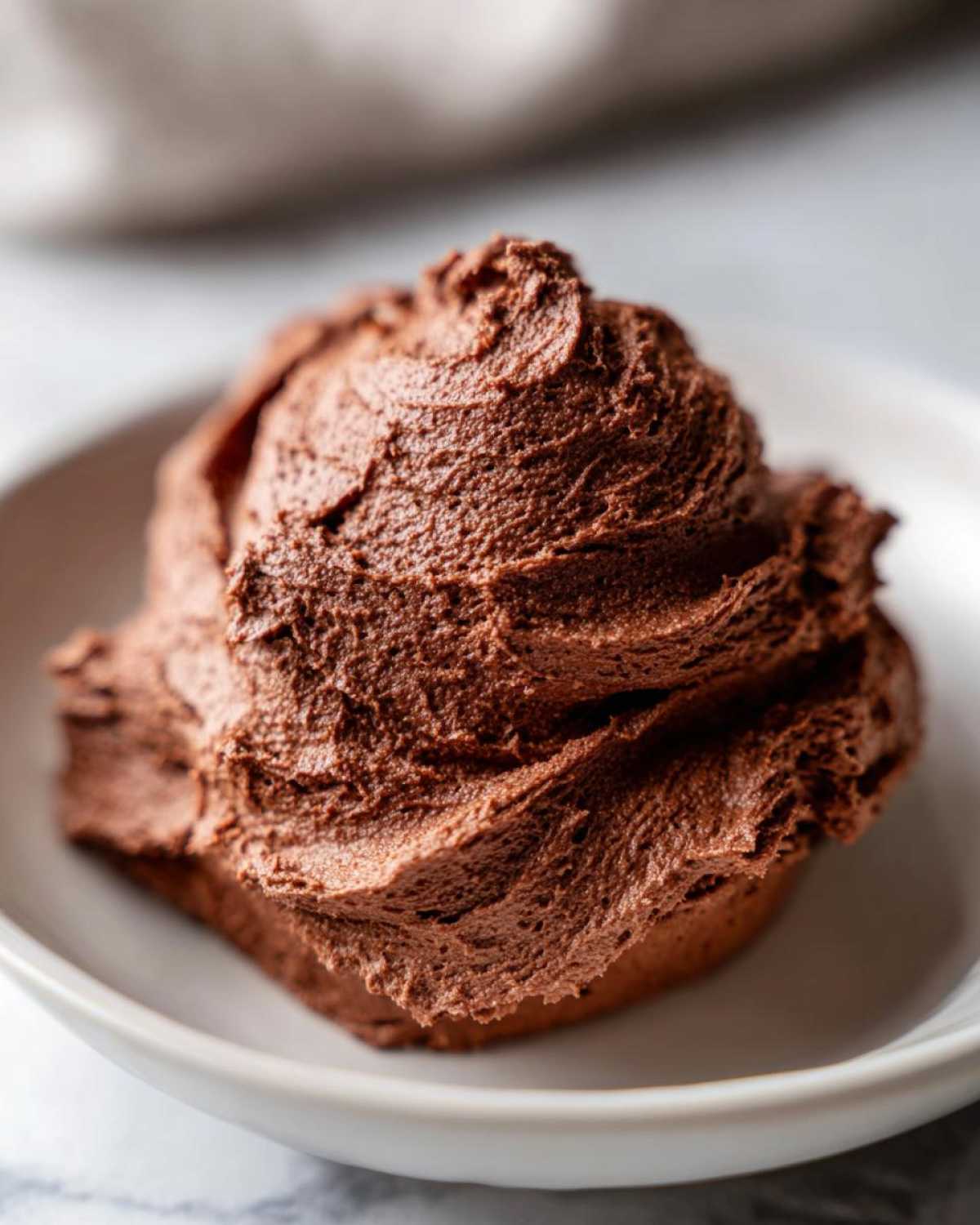 A generous, perfectly swirled scoop of rich, dark brown chocolate frosting in a small white bowl.