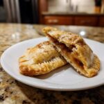 Two golden baked apple pie cookies, one cut open to show the spiced apple filling, dusted with cinnamon.