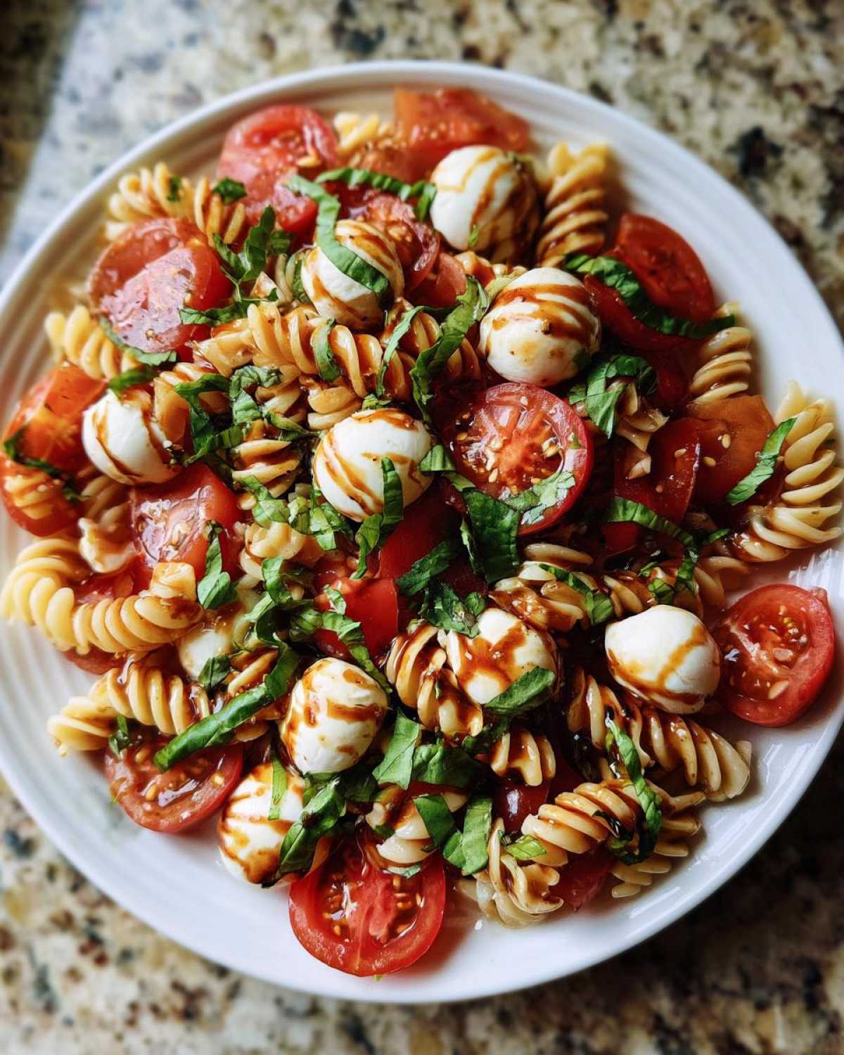 A vibrant bowl of caprese pasta salad featuring fusilli, cherry tomatoes, mozzarella balls, and fresh basil drizzled with balsamic glaze.