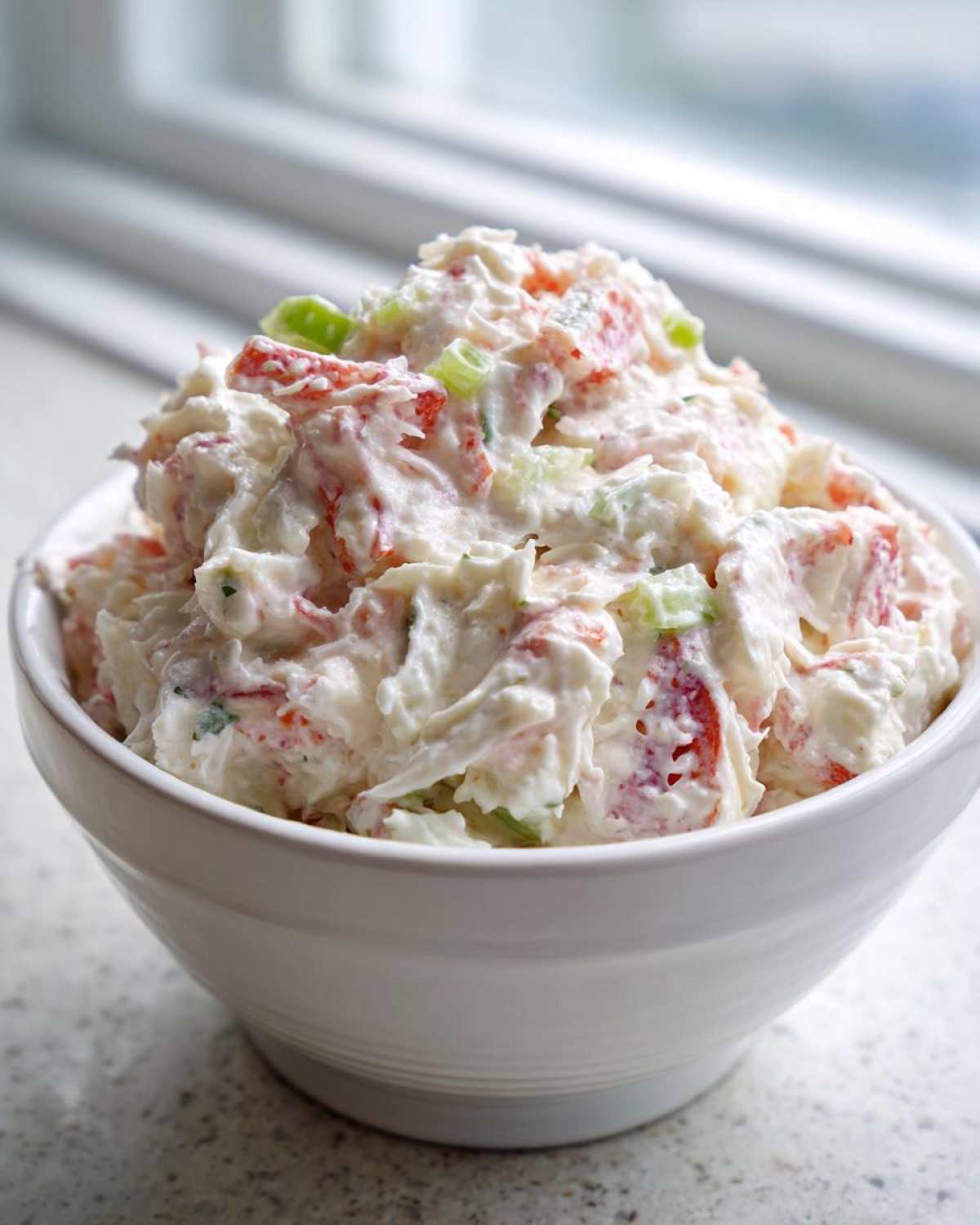 A close-up of creamy, homemade crab salad mixed with red and green ingredients, served in a white bowl.
