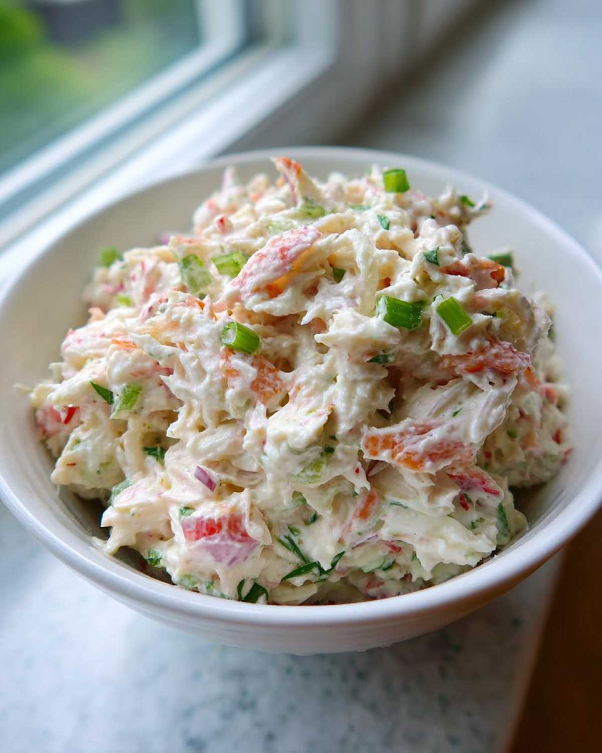 A close-up of creamy crab salad mixed with red peppers and green onions, served in a white bowl.