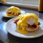 Close-up of delicious eggs benedict topped with bright yellow hollandaise sauce and bacon on an English muffin.