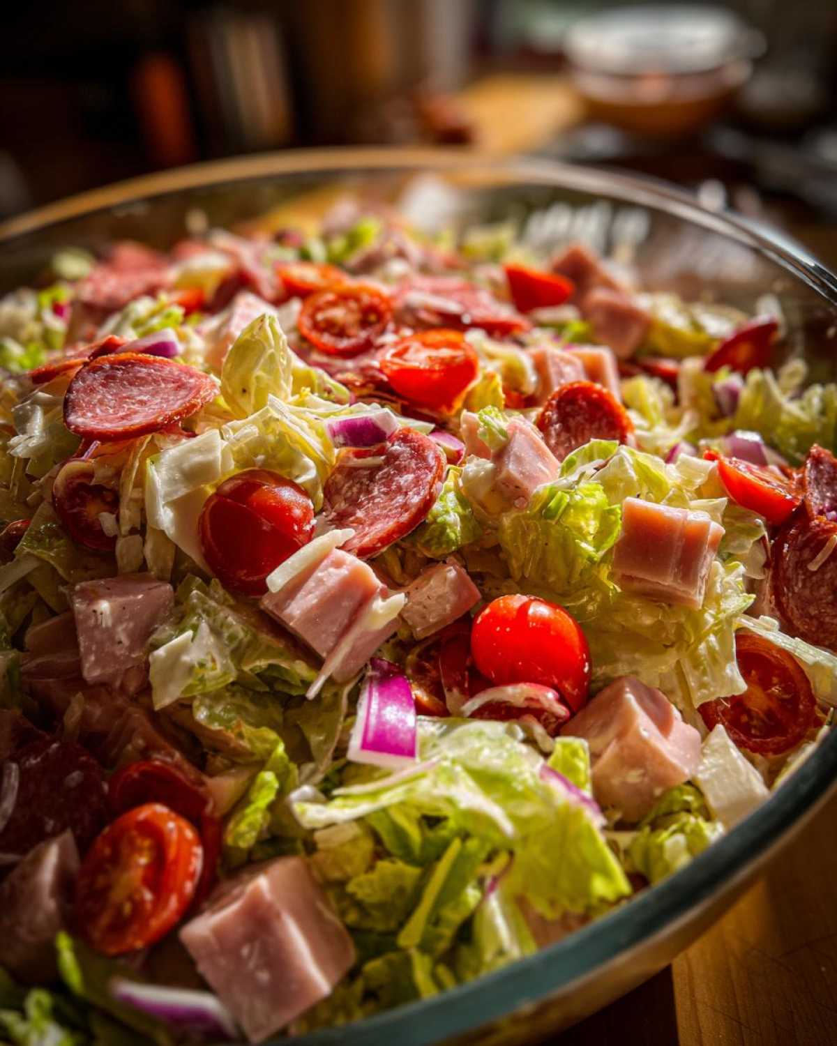 Close-up of a large glass bowl filled with a vibrant grinder salad featuring lettuce, salami, ham cubes, cherry tomatoes, and red onion.