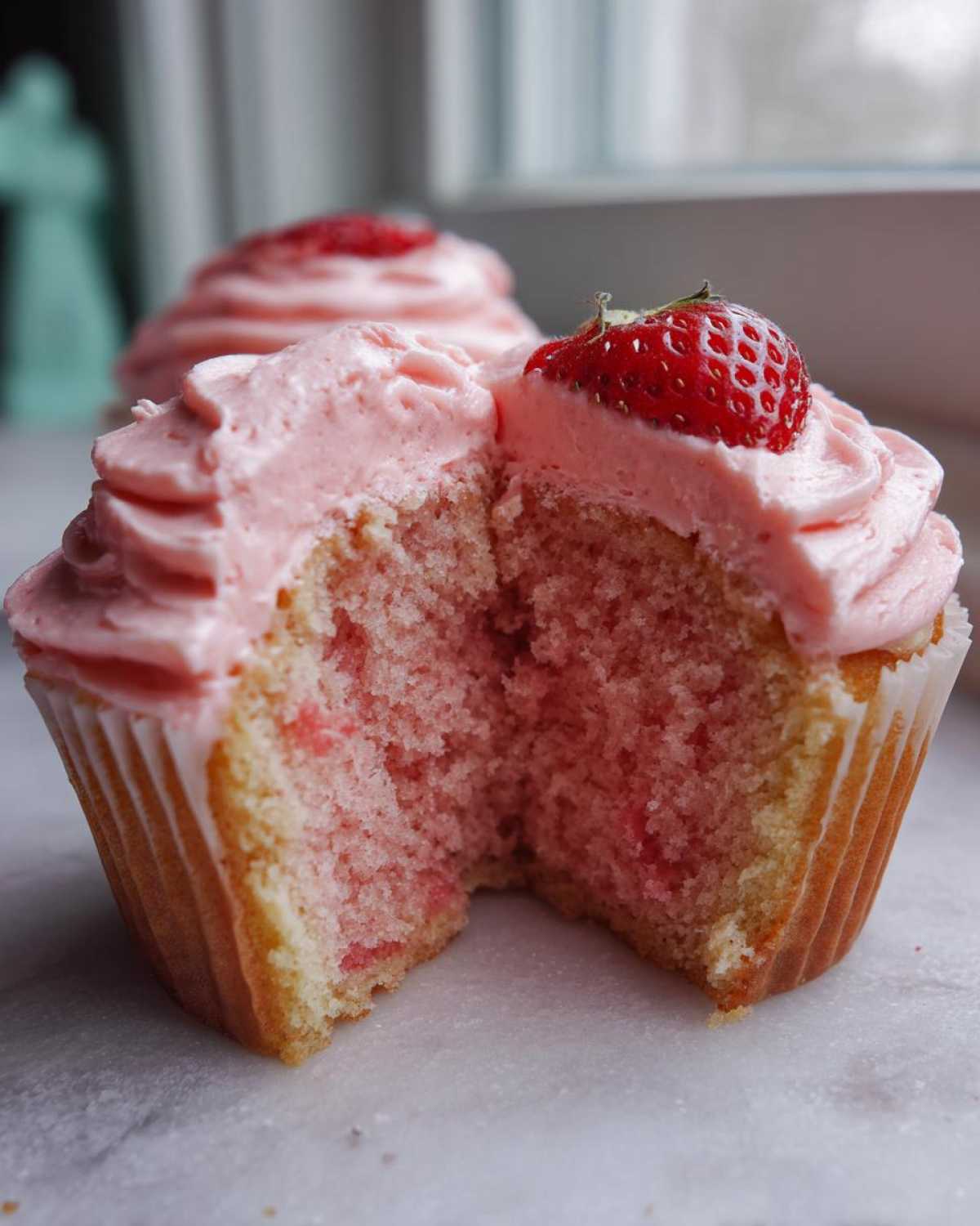A close-up of amazing strawberry cupcakes cut in half, showing the pink crumb and strawberry frosting topped with a fresh strawberry.
