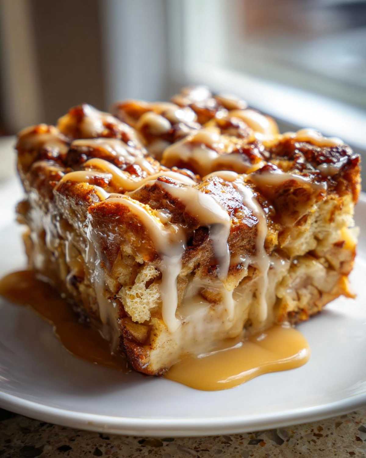 A rich, square slice of apple bread pudding topped with vanilla glaze and caramel sauce.