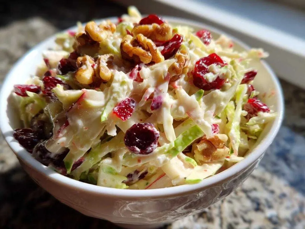 A close-up of creamy apple cranberry coleslaw topped with walnuts and dried cranberries in a white bowl.