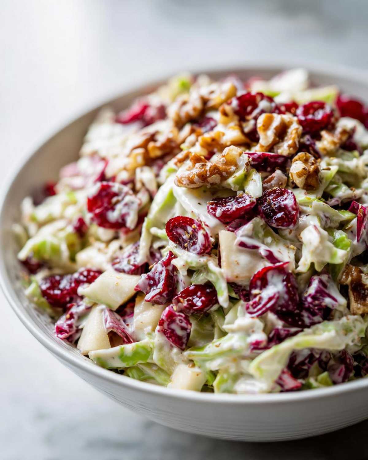 A close-up of creamy apple cranberry coleslaw topped with walnuts and dried cranberries in a white bowl.