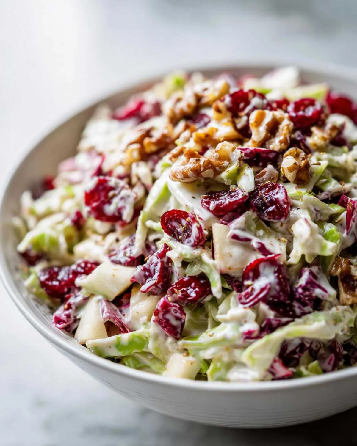 A close-up of creamy apple cranberry coleslaw topped with walnuts and dried cranberries in a white bowl.