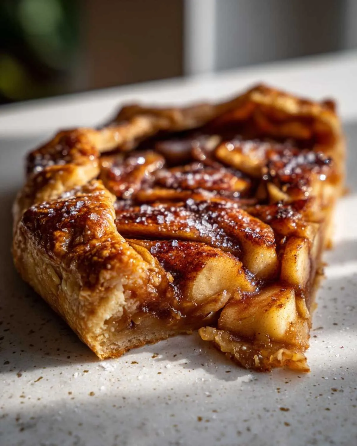 A close-up, sunlit shot of a slice of homemade apple galette showing caramelized apples and flaky crust sprinkled with coarse sugar.
