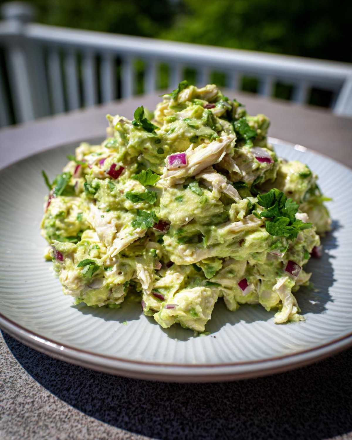 A mound of creamy avocado chicken salad mixed with shredded chicken, red onion, and cilantro, served on a light blue plate outdoors.