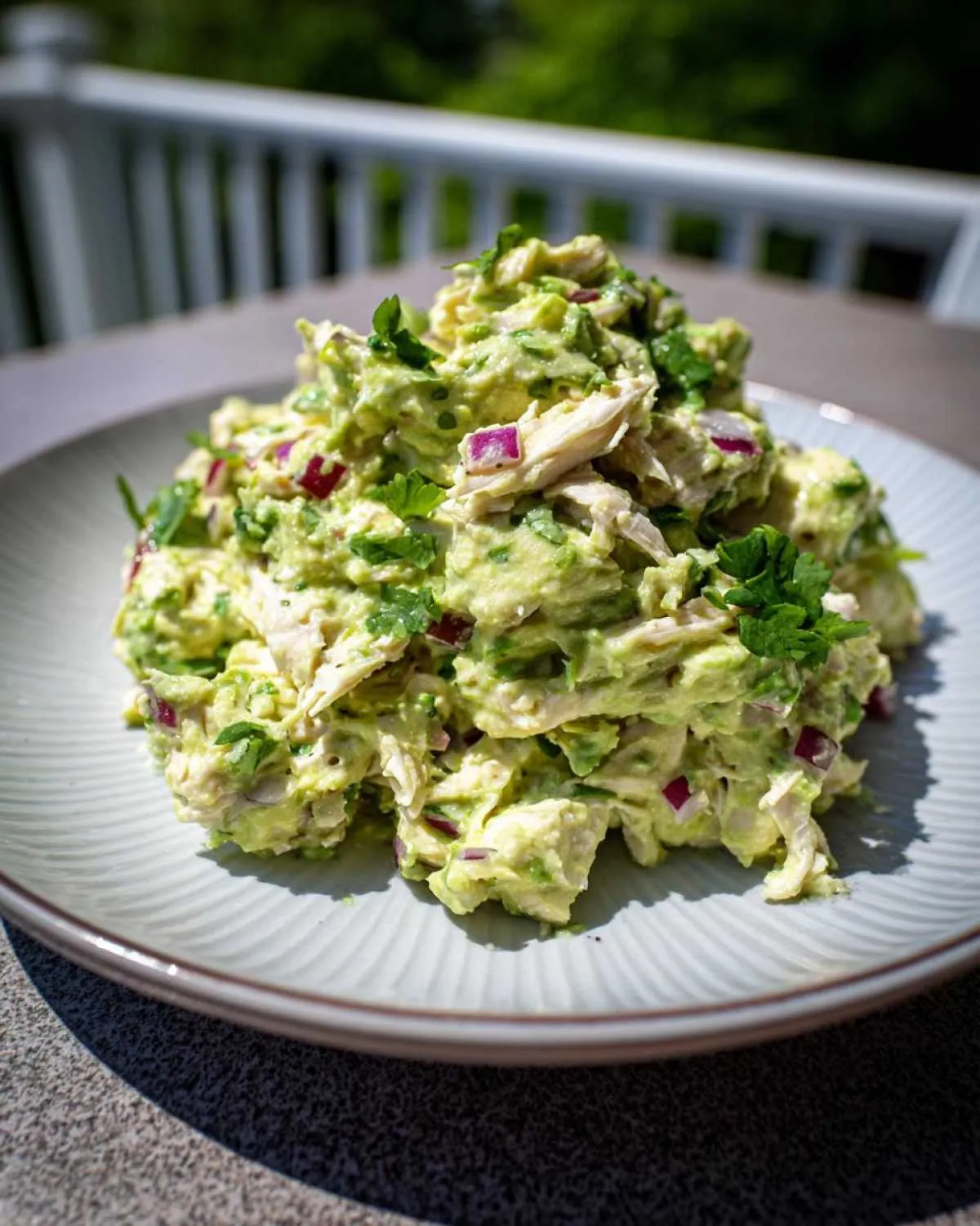 A mound of creamy avocado chicken salad mixed with shredded chicken, red onion, and cilantro, served on a light blue plate outdoors.