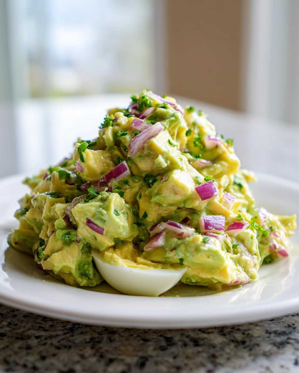 A mound of chunky avocado egg salad mixed with diced red onion and parsley, served with a hard-boiled egg half.