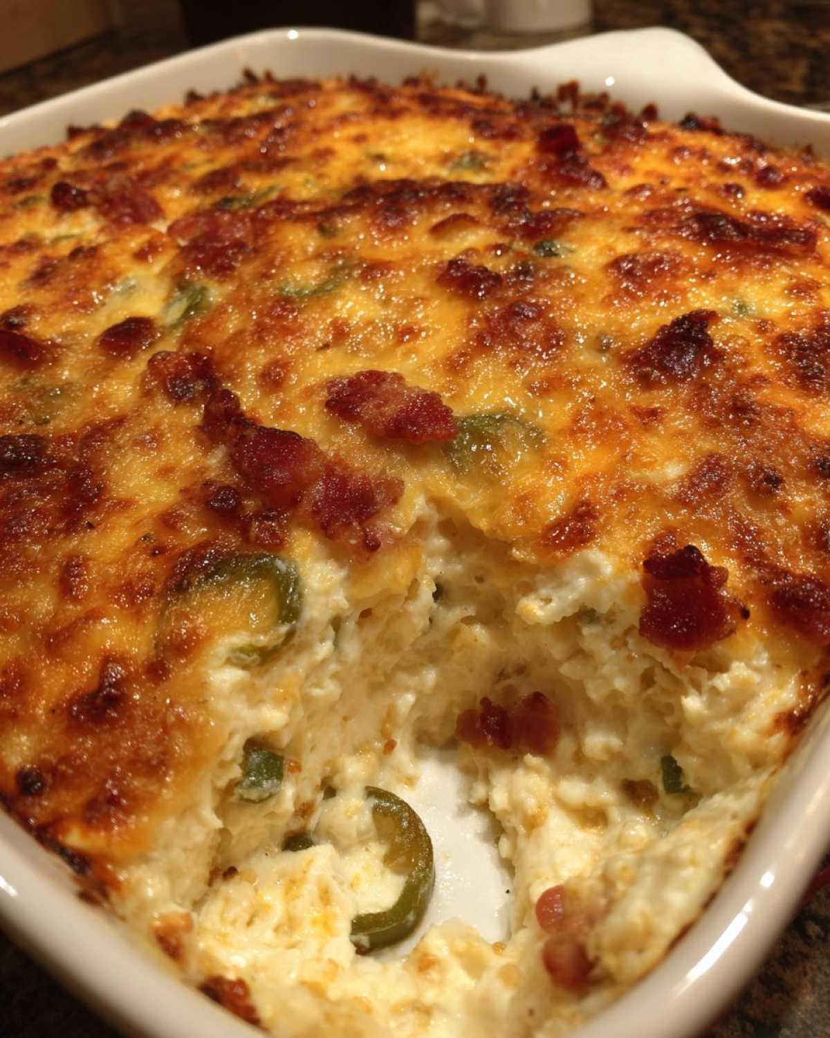 Close-up of a hot, baked popper dip in a white dish with a scoop removed, showing creamy filling, bacon, and jalapeños.