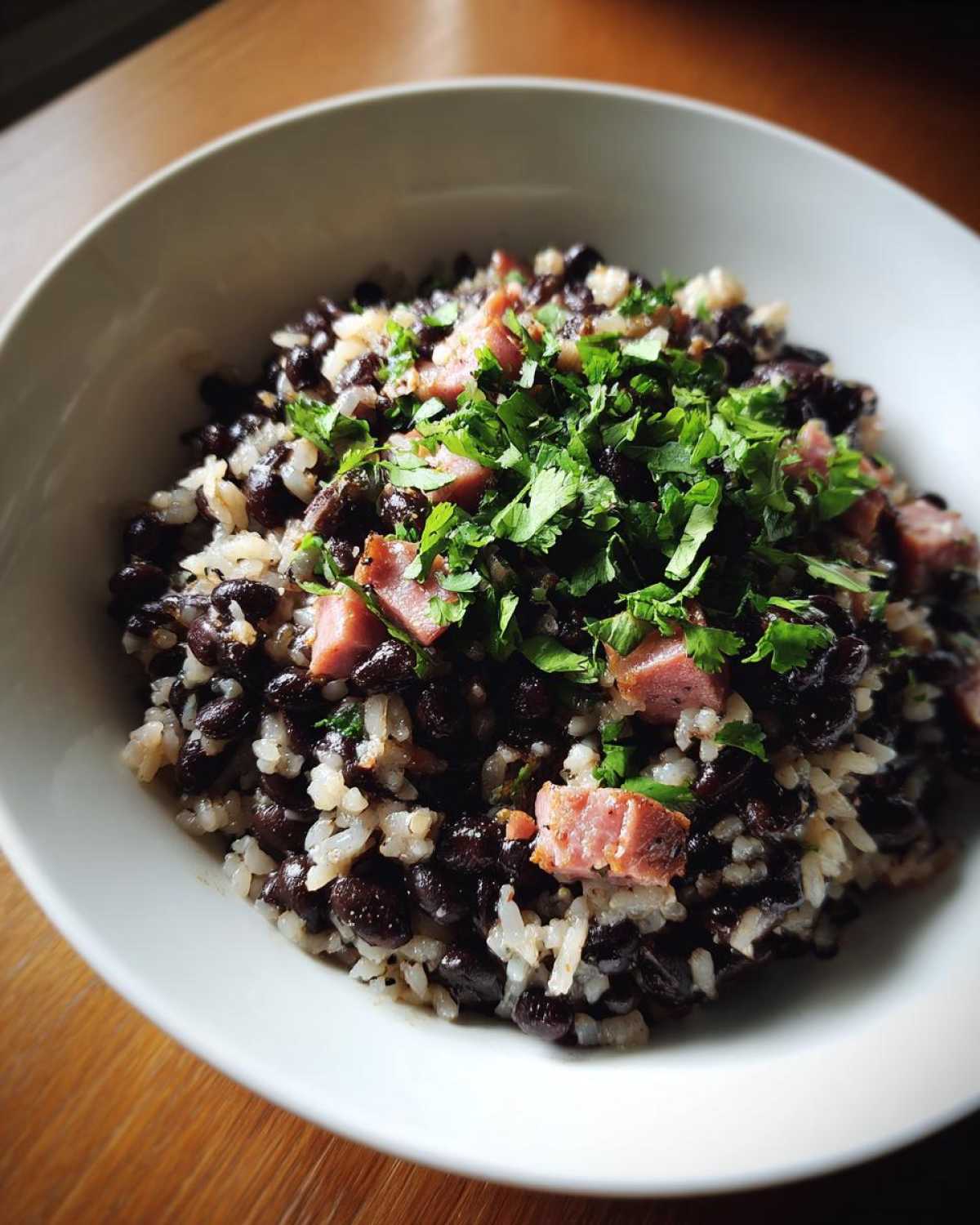 A close-up of a white bowl filled with black beans and rice, topped with diced ham and fresh cilantro.