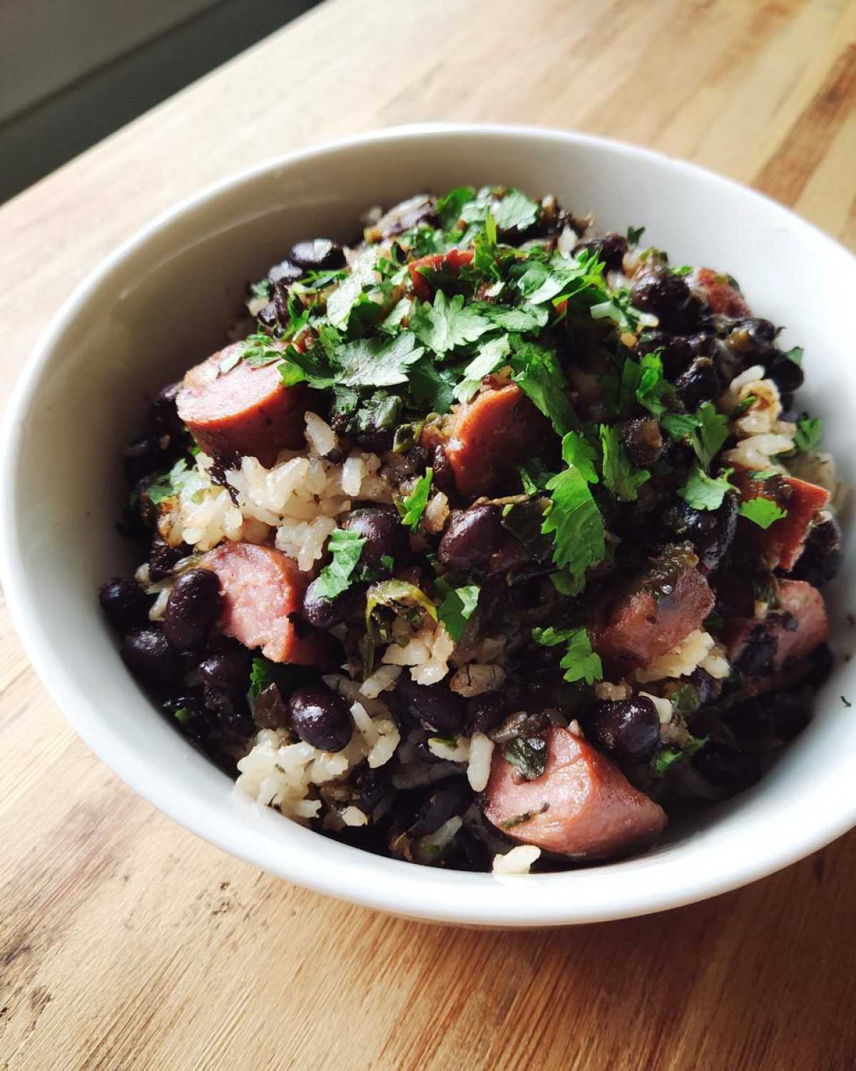 Close-up of a white bowl filled with savory black beans and rice mixed with sliced sausage and topped with fresh cilantro.