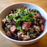 Close-up of a white bowl filled with savory black beans and rice mixed with slices of smoked sausage and topped with fresh cilantro.