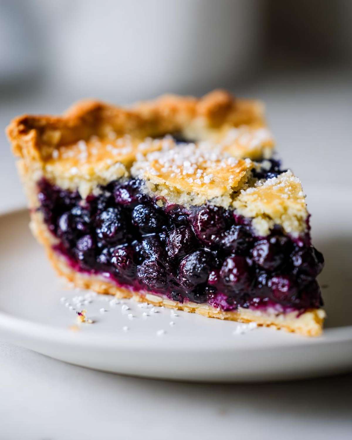 Close-up of a thick slice of blueberry pie recipe showing rich, dark filling and a crumbly top crust dusted with sugar.