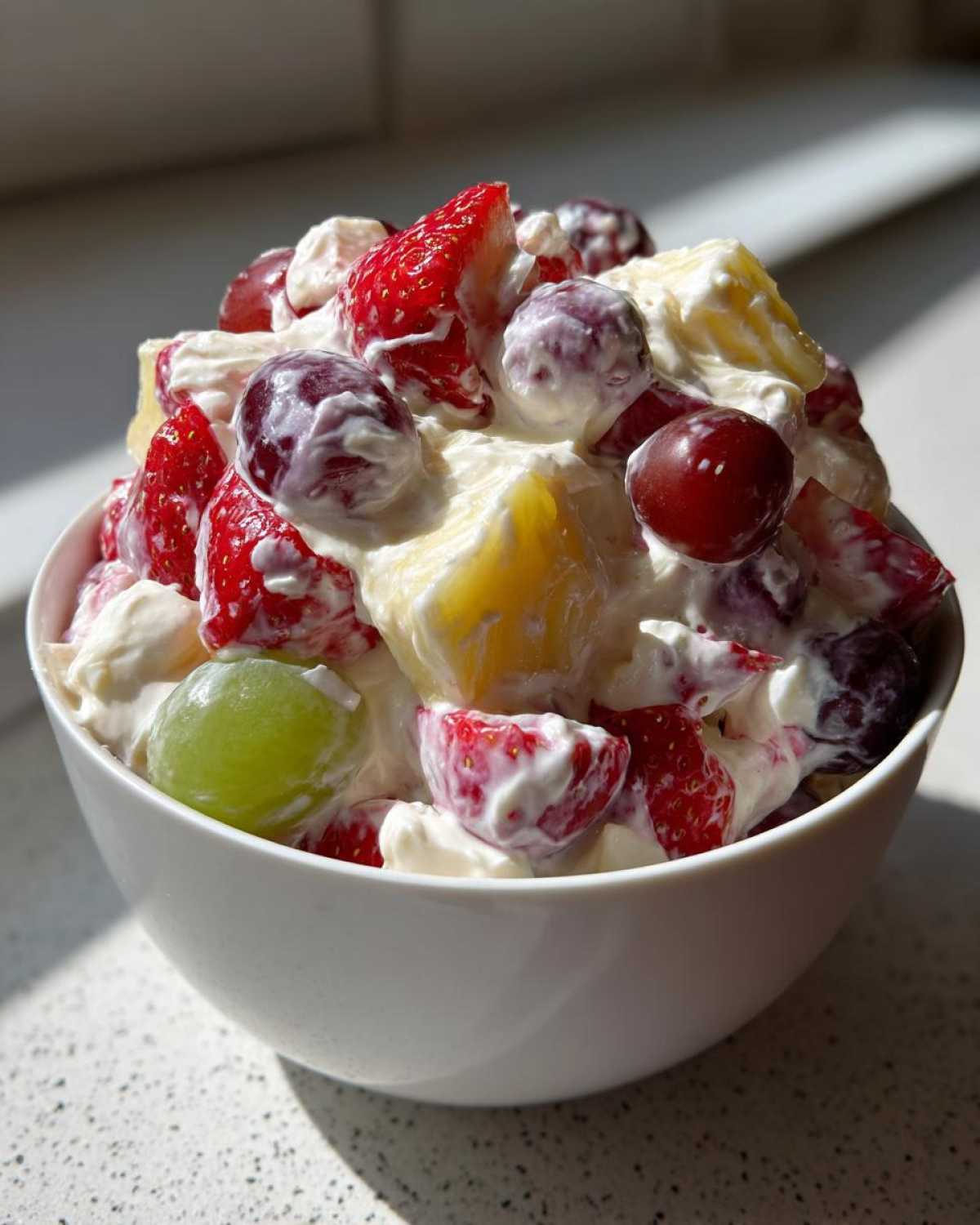 A white bowl filled with creamy cheesecake fruit salad featuring strawberries, red and green grapes, and pineapple chunks.