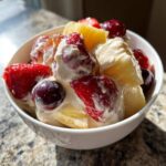 A close-up of a creamy cheesecake fruit salad featuring strawberries, pineapple chunks, and dark cherries in a white bowl.
