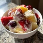 A close-up of a creamy cheesecake fruit salad featuring strawberries, pineapple chunks, and dark cherries in a white bowl.