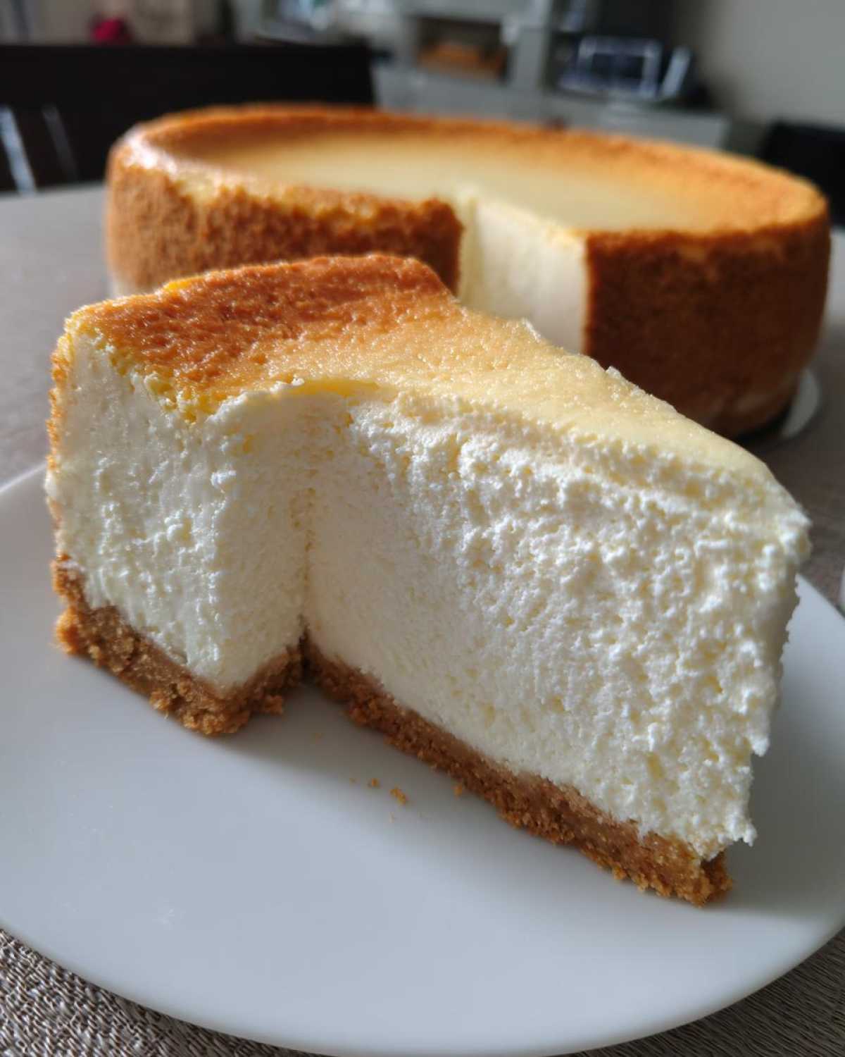 A thick slice of creamy cheesecake with a golden-brown top and graham cracker crust, next to the rest of the cheesecake recipe.