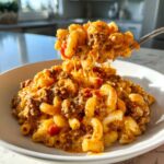 A spoonful of creamy, cheesy taco pasta being lifted from a white bowl, showing ground beef and elbow macaroni.