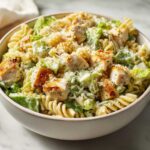 A creamy bowl of chicken caesar pasta salad featuring rotini pasta, grilled chicken chunks, and romaine lettuce.