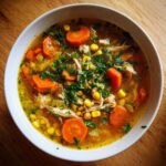 Close-up of a bowl of rich chicken vegetable soup, featuring shredded chicken, bright orange carrots, corn, and fresh parsley garnish.
