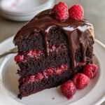 A decadent slice of chocolate raspberry cake layered with jam, topped with chocolate ganache and fresh raspberries.
