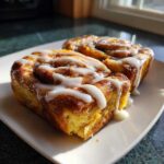 Two servings of cinnamon roll french toast smothered in white icing, sitting on a white plate.