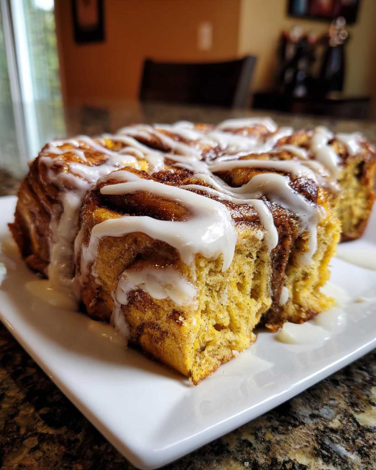 A close-up of a serving of cinnamon roll french toast drenched in thick white icing on a white plate.