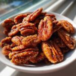 A close-up shot of glossy, caramelized cinnamon sugar pecans piled high in a small white bowl, catching the sunlight.