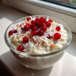 A glass bowl of creamy coconut chia pudding topped with fresh red currants and shredded coconut.