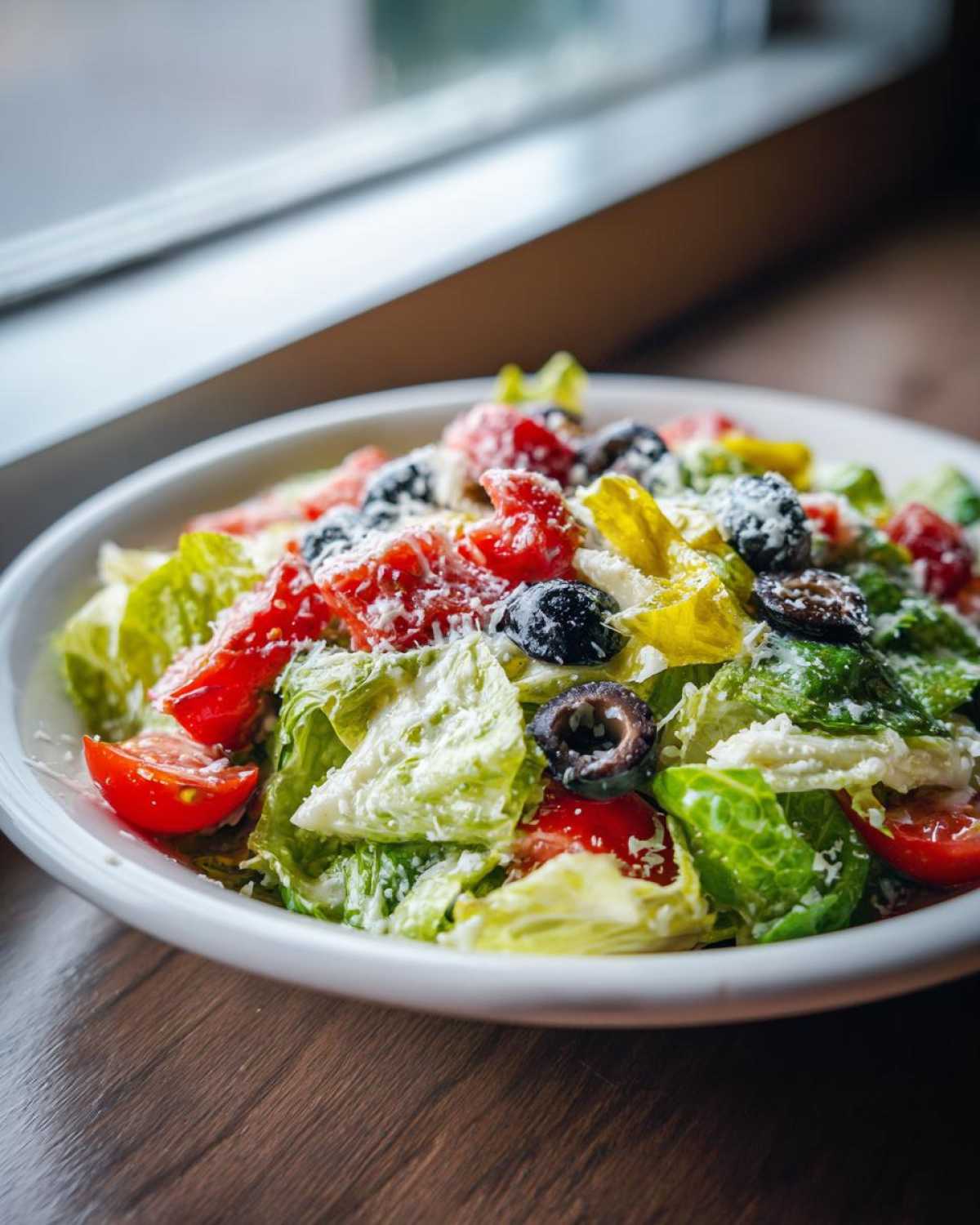 A close-up of a bowl filled with crisp lettuce, tomatoes, black olives, and pepperoncini, topped with grated cheese, resembling a copycat Olive Garden salad.