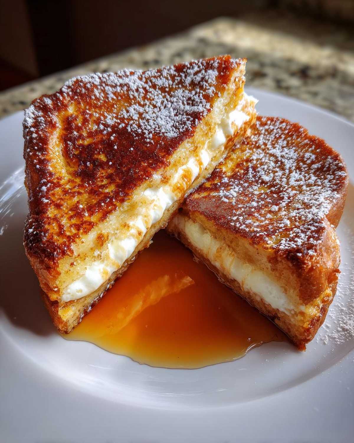 Two halves of golden brown stuffed french toast filled with cream cheese, dusted with powdered sugar, and sitting in maple syrup.
